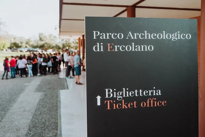 Un cartel que dice Parco Archeologico di Ercolano, Biglietteria, Ticket office, con un grupo haciendo cola detrás